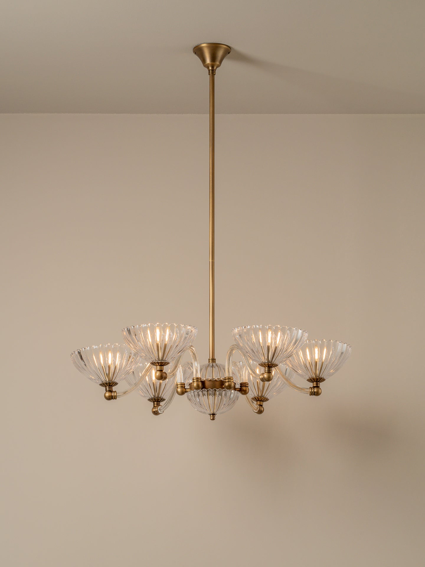 A six-light brass chandelier with clear ribbed glass shades, suspended from a matching brass ceiling canopy against a neutral beige wall.