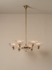 A six-light brass chandelier with clear ribbed glass shades, suspended from a matching brass ceiling canopy against a neutral beige wall.