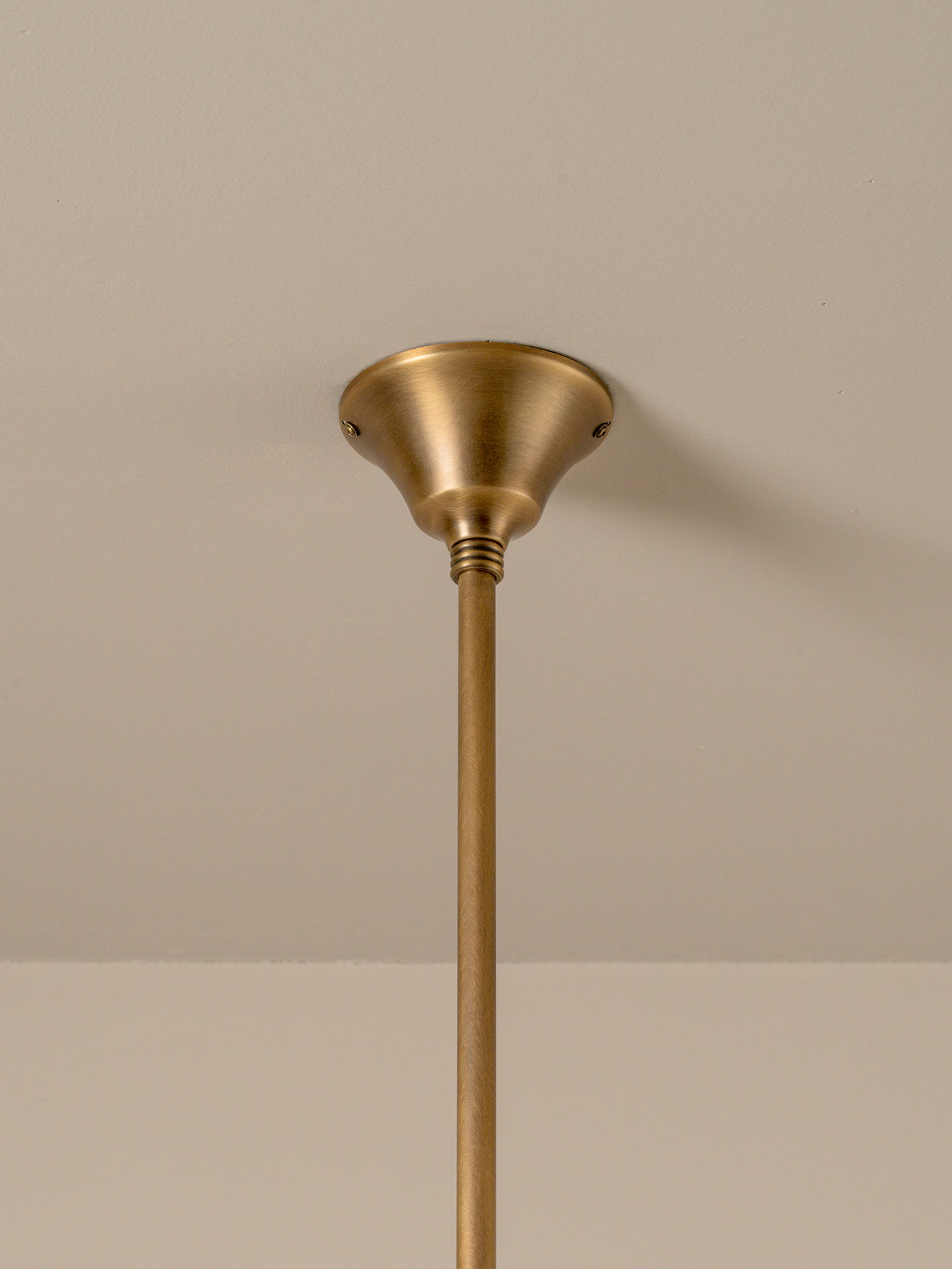 A close-up of a brass ceiling light fixture showing the round ceiling canopy and the top of the downrod against a neutral beige wall.