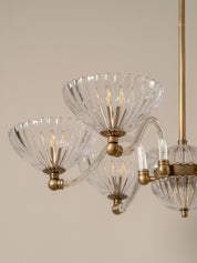 A close-up of a multi-light chandelier with clear ribbed glass shades and brushed brass metal arms and finials, shown against a plain beige background.