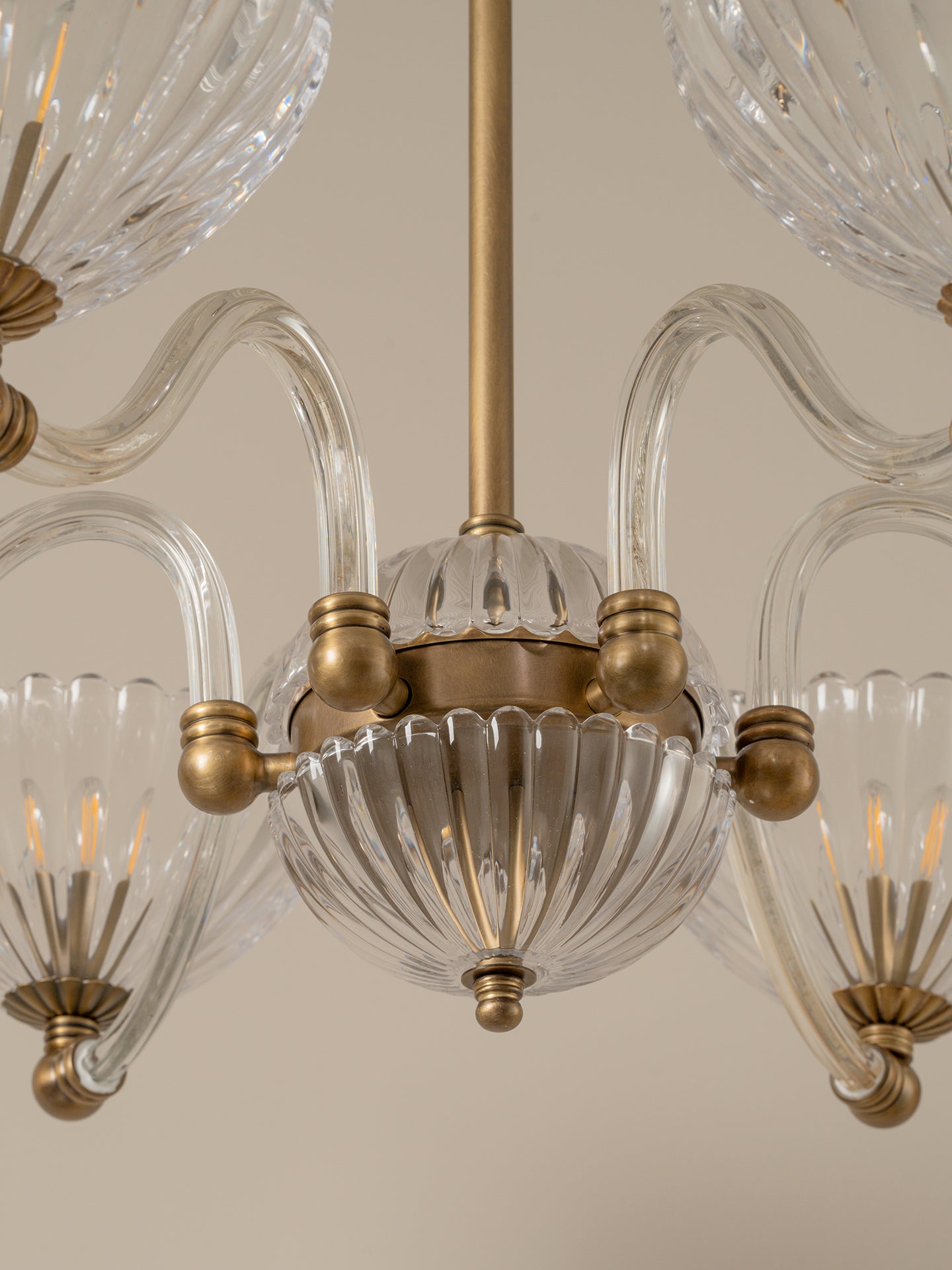 A close-up of a multi-light chandelier featuring clear ribbed glass shades and curved clear glass arms with antique brass hardware, shown against a plain beige background.