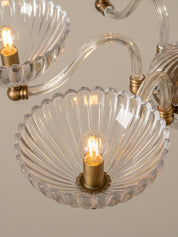 A close-up of a clear ribbed glass wall sconce with a warm glowing bulb and brass metal hardware, shown against a soft beige background.