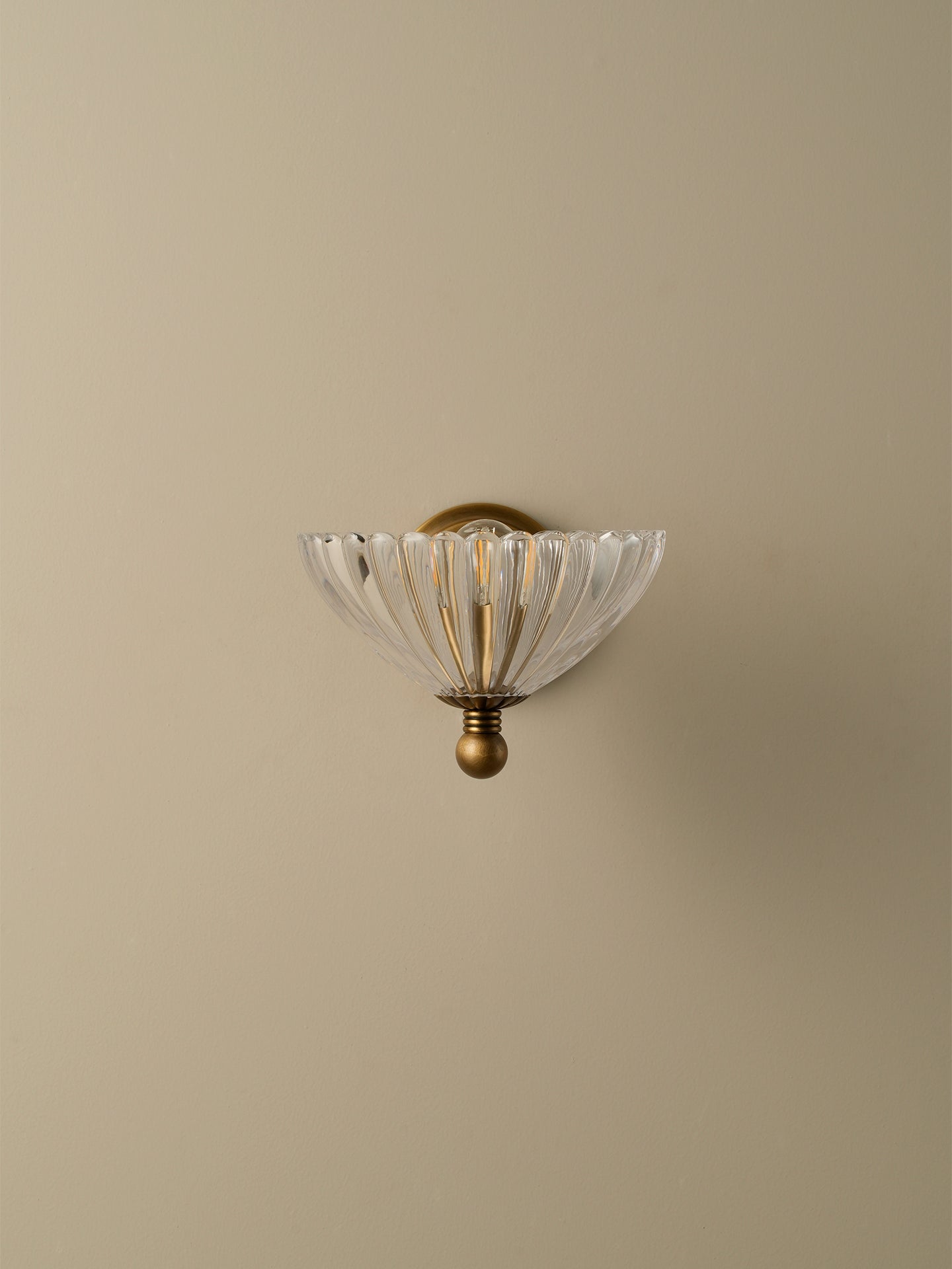 A vintage-style wall sconce with a clear ribbed glass shade and an antique brass/bronze metal mount, shown against a solid beige wall.