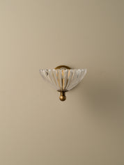 A vintage-style wall sconce with a clear ribbed glass shade and an antique brass/bronze metal mount, shown against a solid beige wall.