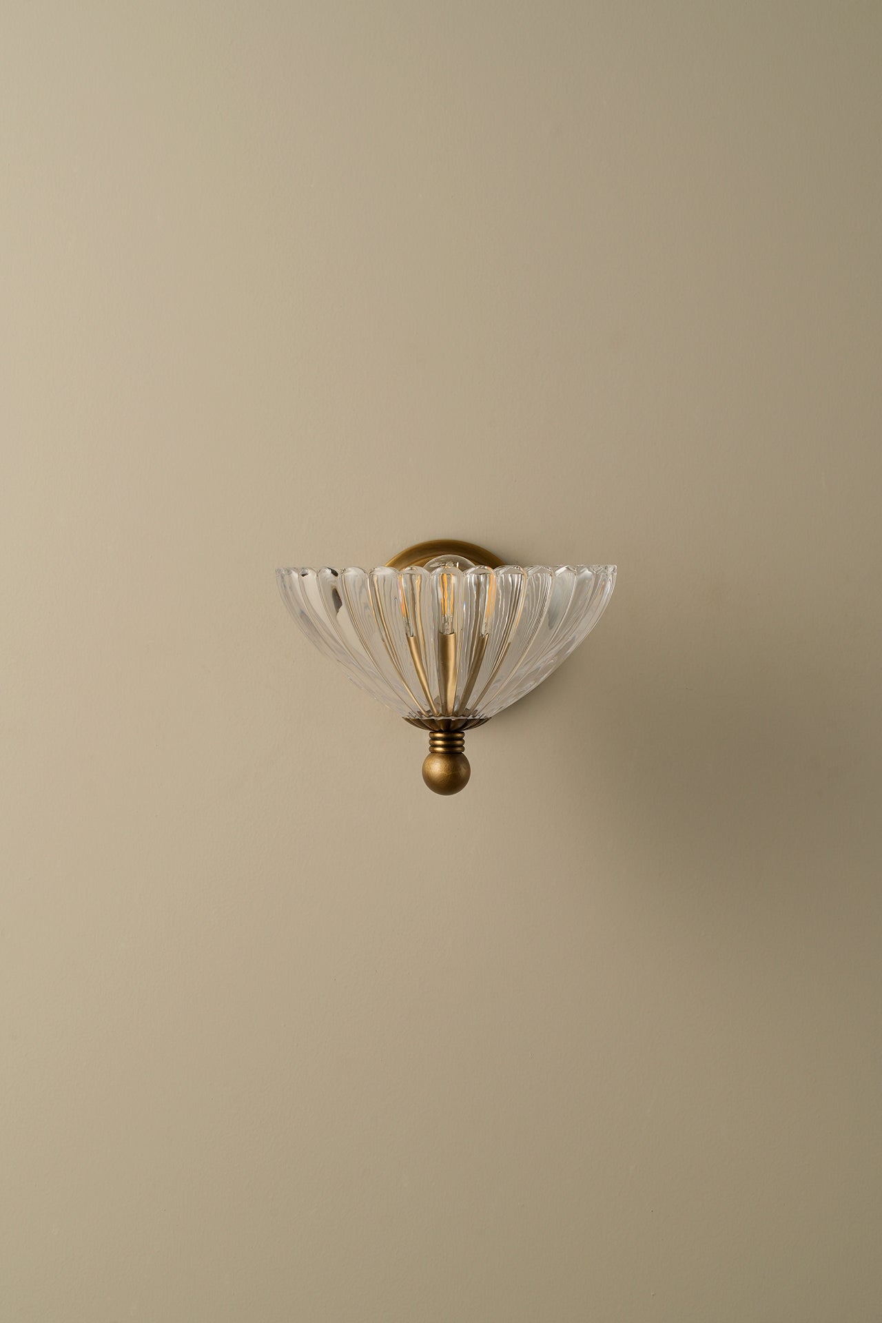 A wall-mounted sconce with a clear ribbed glass shade and an antique brass or bronze metal fixture, shown against a plain beige wall.