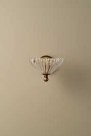 A wall-mounted sconce with a clear ribbed glass shade and an antique brass or bronze metal fixture, shown against a plain beige wall.