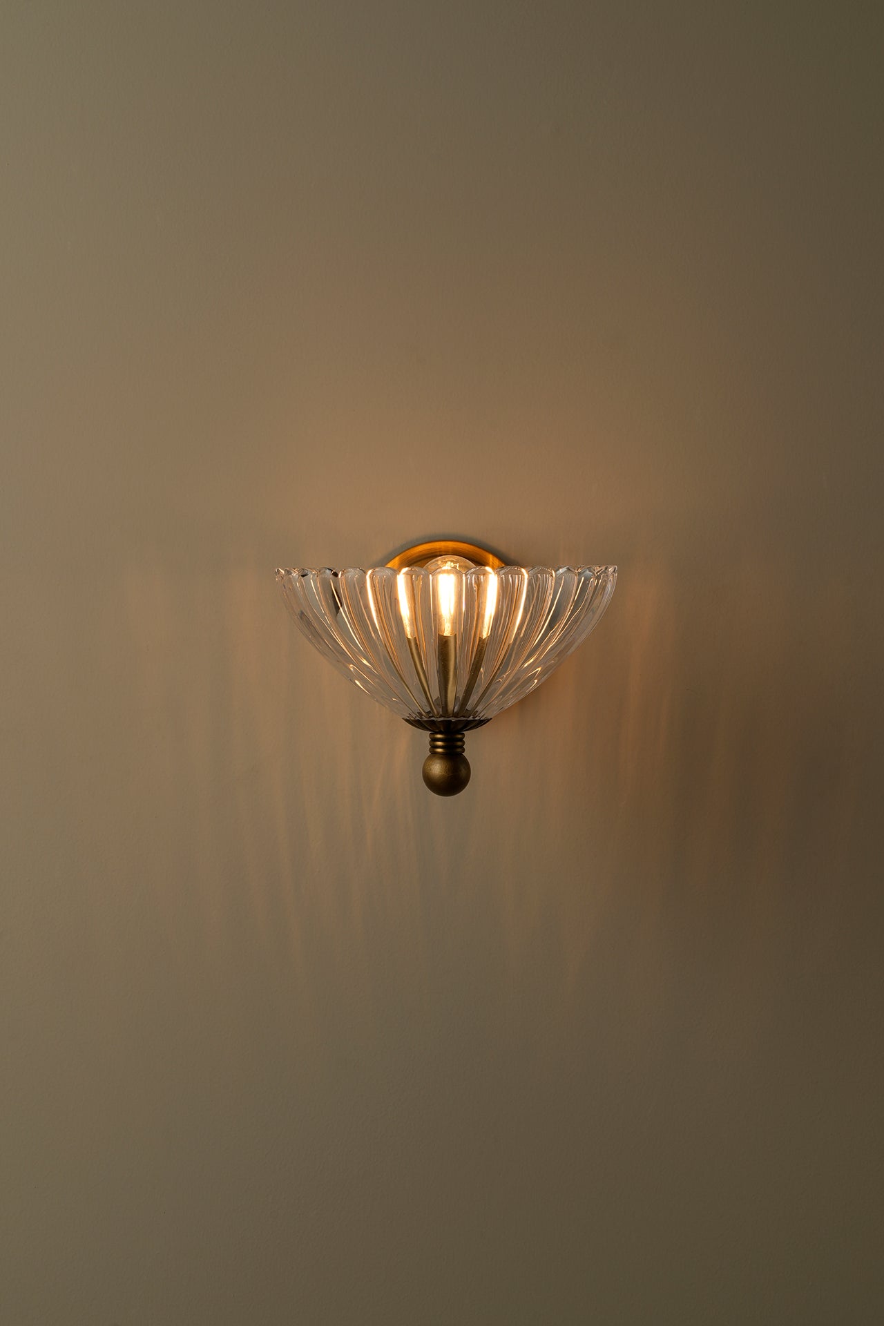 A wall-mounted sconce with a clear ribbed glass shade and an antique bronze metal frame, illuminated against a warm beige wall.