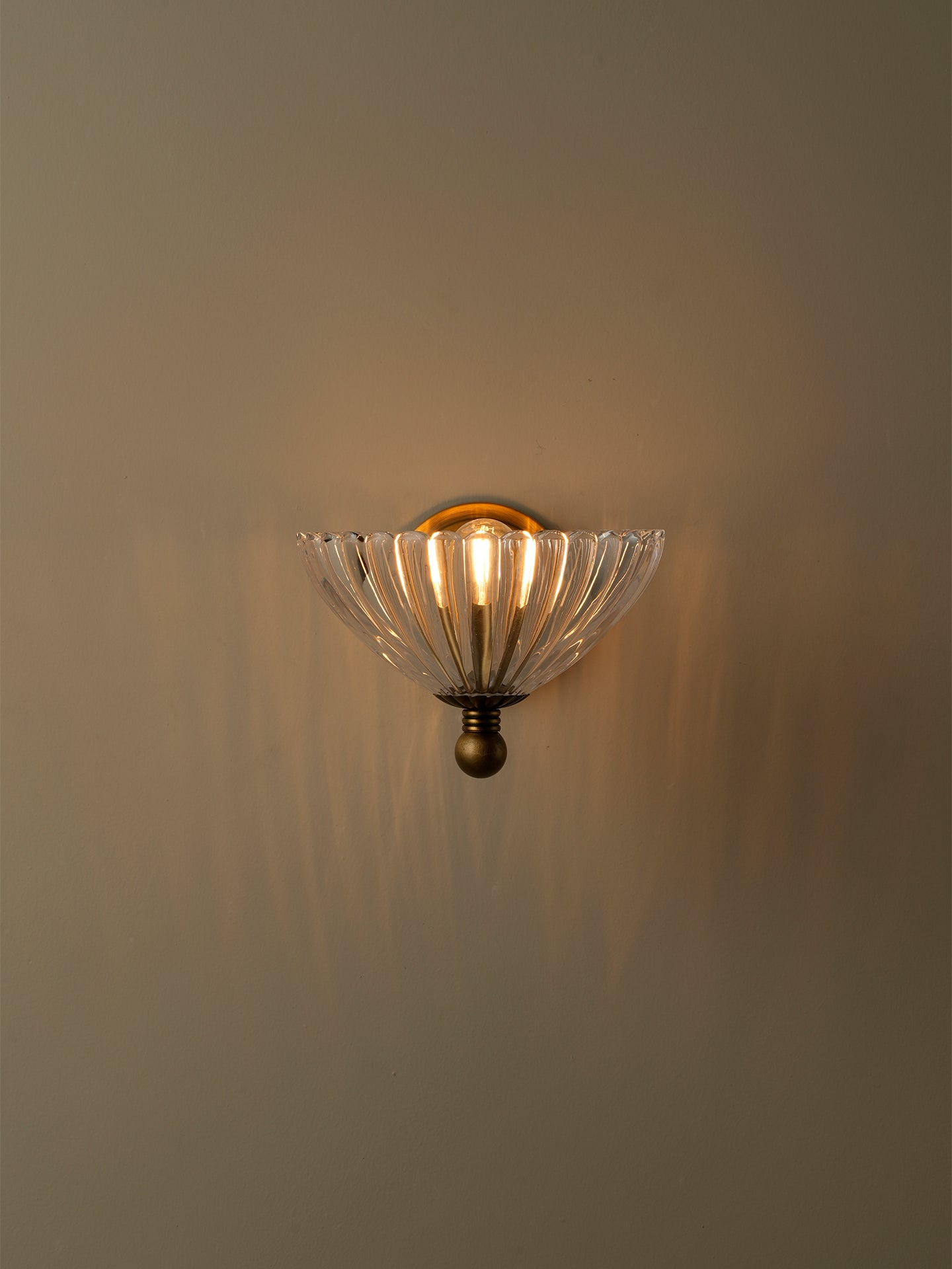 A wall-mounted sconce with a clear ribbed glass shade and an antique bronze metal base, shown lit against a warm beige wall.
