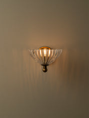 A wall-mounted sconce with a clear ribbed glass shade and an antique bronze metal base, shown lit against a warm beige wall.