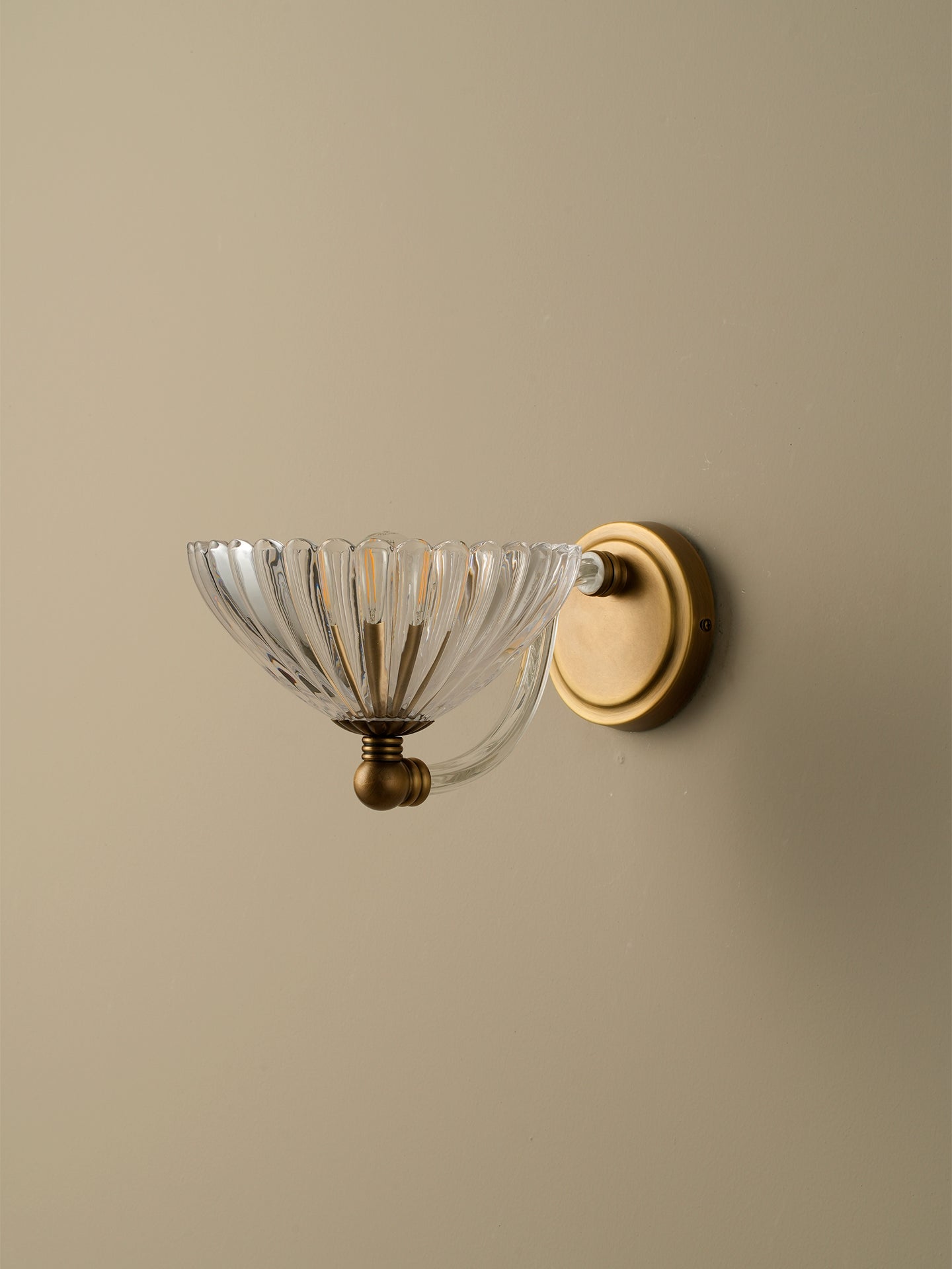 A wall-mounted sconce with a clear ribbed glass bowl shade and a round brass backplate, mounted on a warm beige wall.