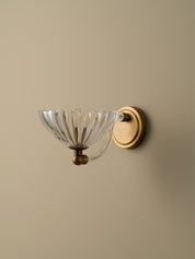 A wall-mounted sconce with a clear ribbed glass bowl shade and a round brass backplate, mounted on a warm beige wall.