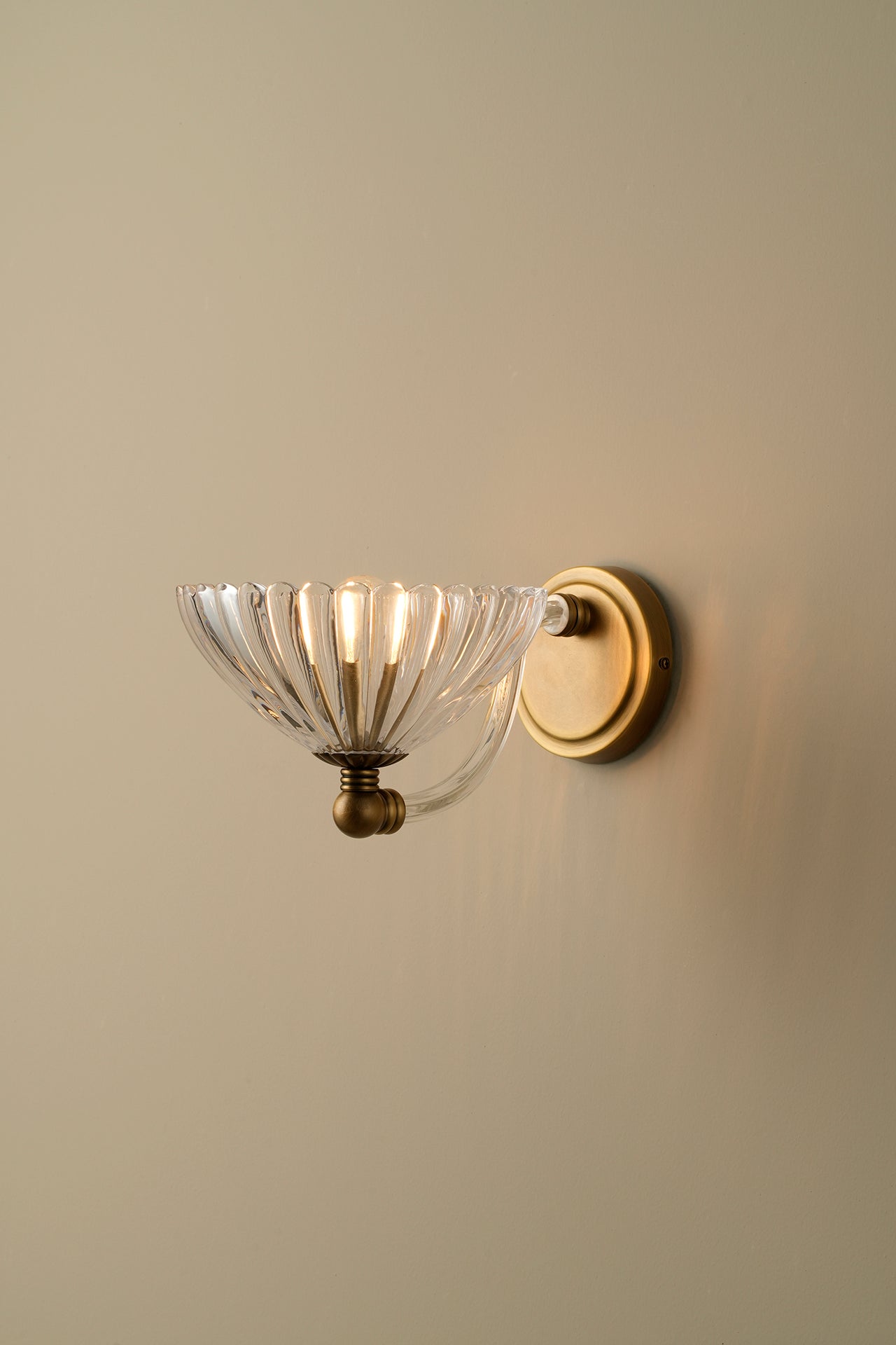 A wall-mounted sconce with a clear ribbed glass bowl shade and a warm brass or gold-toned metal mount, shown against a plain beige wall.
