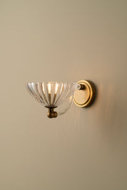 A wall-mounted sconce with a clear ribbed glass bowl shade and a warm brass or gold-toned metal mount, shown against a plain beige wall.