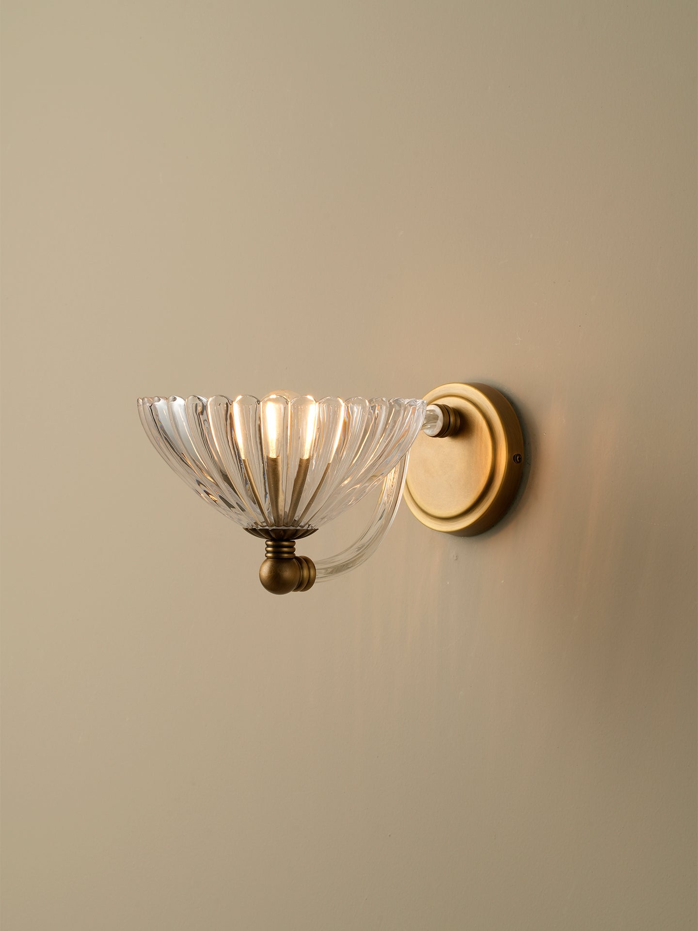 A wall-mounted sconce with a clear ribbed glass bowl shade and an antique brass or bronze metal base and backplate, shown lit against a plain beige wall.