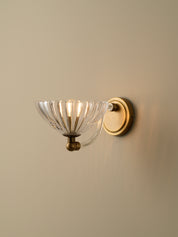 A wall-mounted sconce with a clear ribbed glass bowl shade and an antique brass or bronze metal base and backplate, shown lit against a plain beige wall.