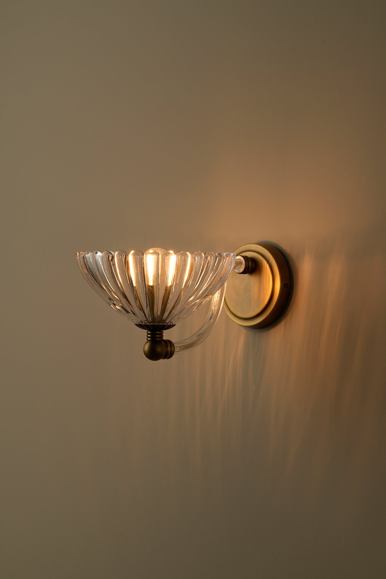 A wall-mounted sconce with a clear ribbed glass bowl shade and a warm brass or bronze metal base, shown lit against a plain beige wall.