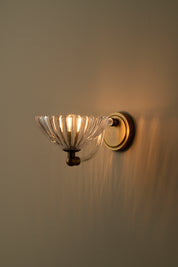 A wall-mounted sconce with a clear ribbed glass bowl shade and a warm brass or bronze metal base, shown lit against a plain beige wall.