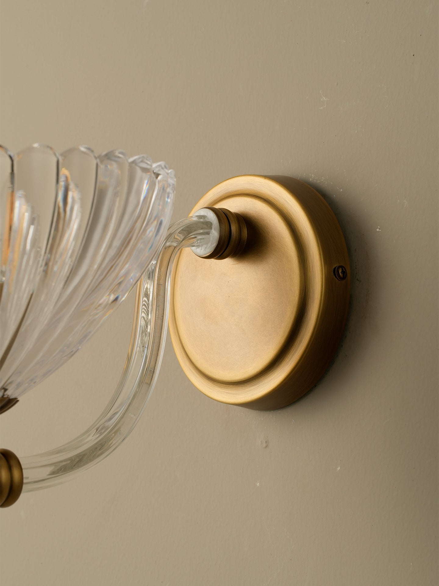 Close-up of a wall-mounted light fixture with a round brushed brass or gold metal backplate and clear ribbed glass arms against a warm beige wall.