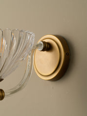 Close-up of a wall-mounted light fixture with a round brushed brass or gold metal backplate and clear ribbed glass arms against a warm beige wall.