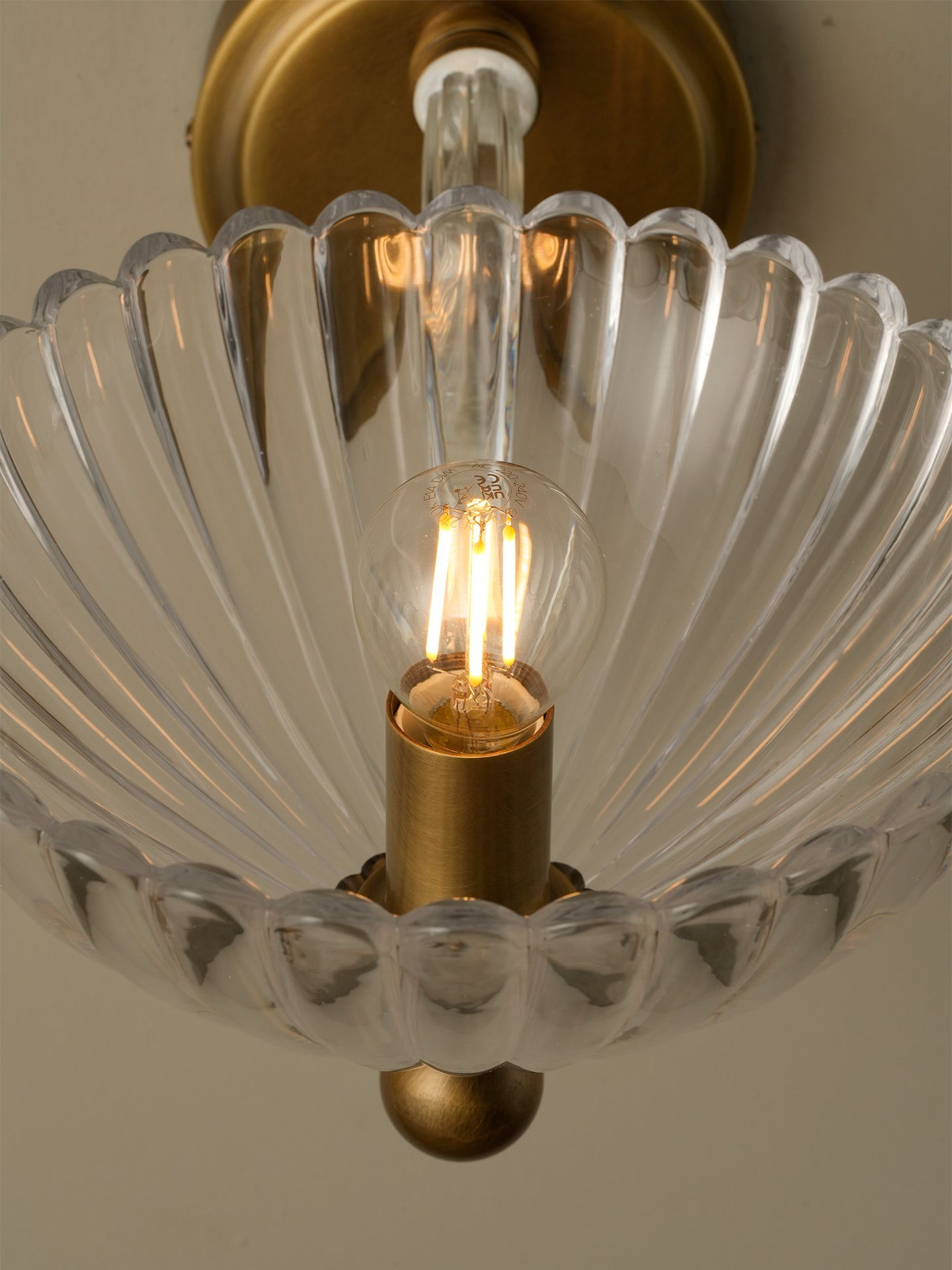A close-up of a vintage-style ceiling light fixture with a clear ribbed glass scallop-shaped shade and brass hardware, illuminated against a neutral background.