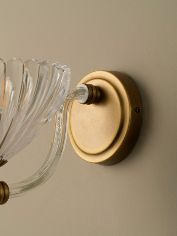 Wall-mounted light fixture with clear glass shade and gold metal base on a beige wall.