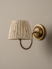 A wall-mounted sconce with a pleated fabric shade featuring a delicate gray botanical pattern, attached to an antique brass curved arm and circular wall plate against a neutral beige wall.
