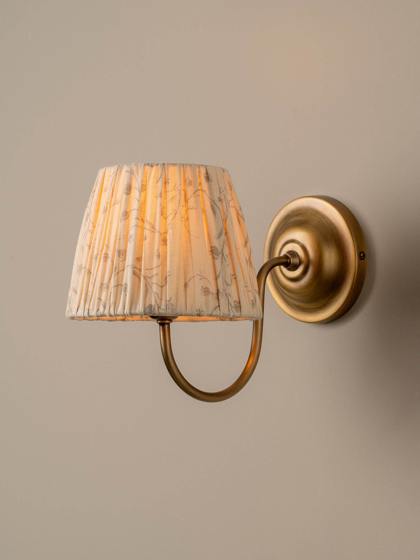 A wall-mounted sconce with a pleated fabric shade in a beige and white floral pattern, illuminated with a warm yellow glow, attached to an antique brass curved arm and circular wall plate against a neutral beige wall.