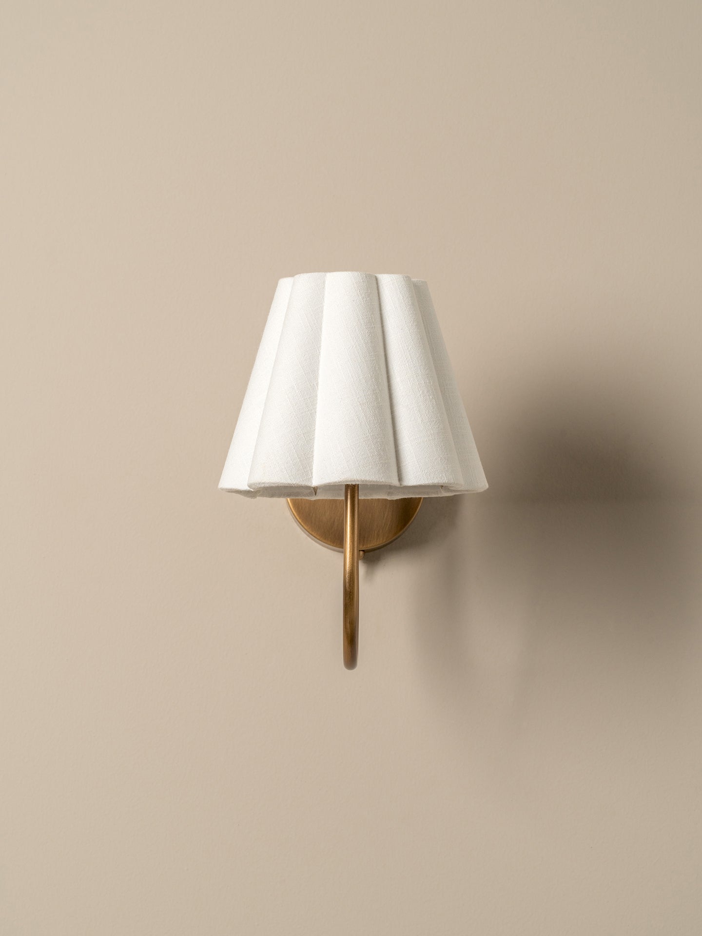 A wall-mounted sconce with a white pleated fabric shade and a brushed brass metal arm, shown against a warm beige wall.