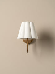 A wall-mounted sconce with a white pleated fabric shade and a brushed brass metal arm, shown against a warm beige wall.