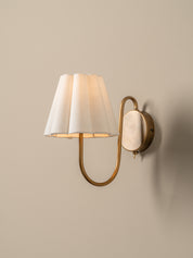 A wall-mounted sconce with a warm brass-toned metal frame and a pleated off-white fabric shade, shown lit against a plain beige wall.