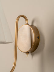 A close-up of a wall-mounted light fixture with a curved brass arm and a round backplate, shown against a warm beige wall with part of a white lampshade visible on the left.
