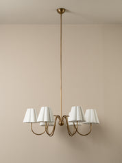 A modern six-light chandelier with a brushed brass or bronze metal frame and white tapered fabric shades, suspended from a matching ceiling canopy against a plain beige wall.