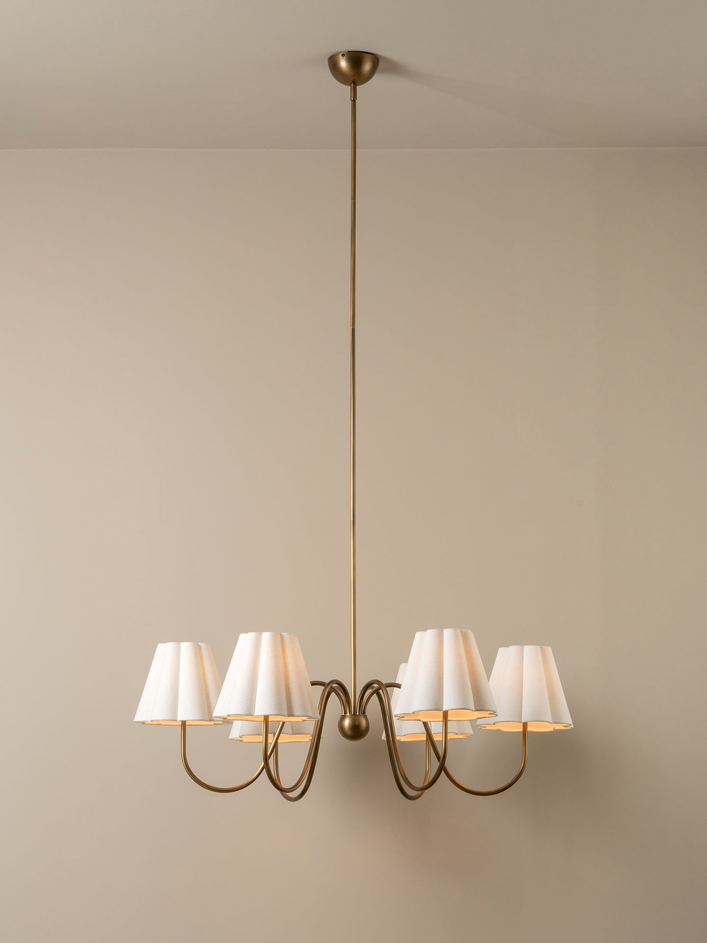 A six-light chandelier with a warm brass-finished metal frame and white pleated fabric shades, suspended from a ceiling against a neutral beige wall.