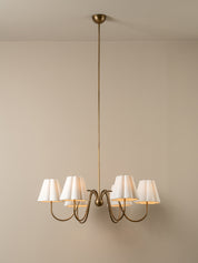 A six-light chandelier with a warm brass-finished metal frame and white pleated fabric shades, suspended from a ceiling against a neutral beige wall.