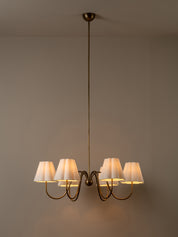 A six-light brass chandelier with white pleated drum shades, suspended from a ceiling against a neutral gray wall.