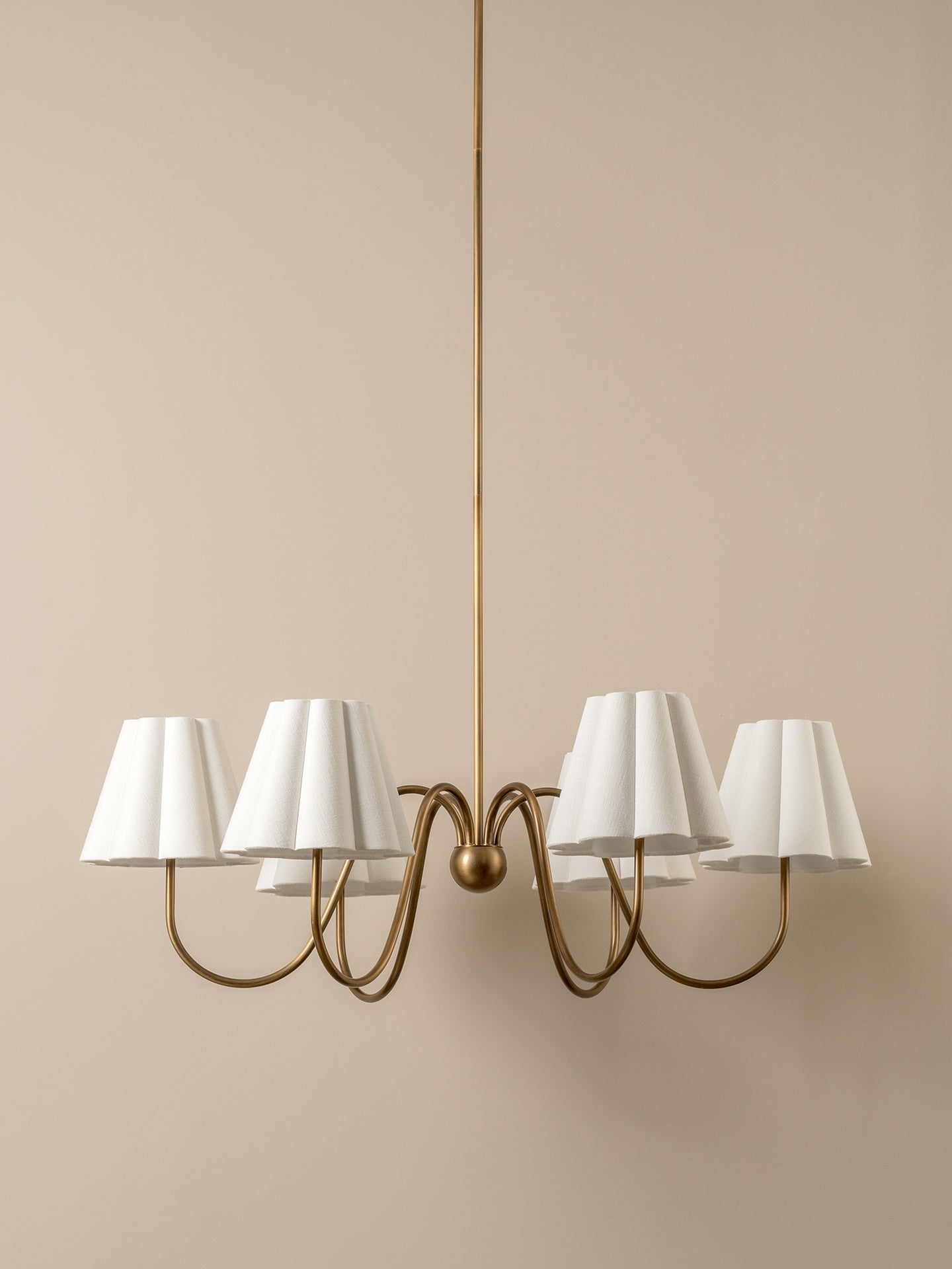 A modern six-light chandelier with a warm brass-finished metal frame and white pleated drum shades, suspended against a solid beige wall.