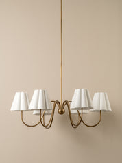 A modern six-light chandelier with a warm brass-finished metal frame and white pleated drum shades, suspended against a solid beige wall.