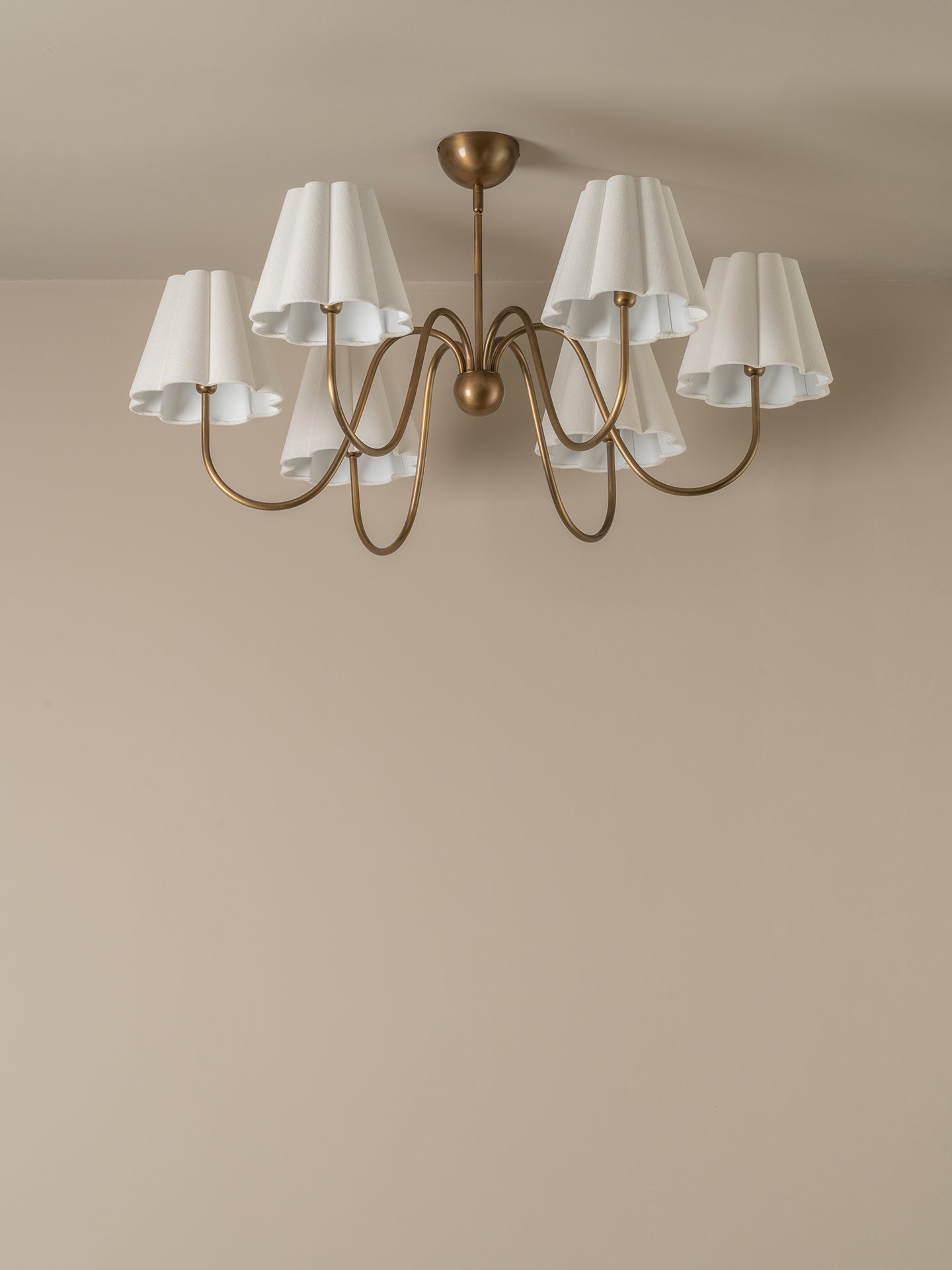 A six-light chandelier with a curved brass-finished metal frame and white pleated drum shades, shown hanging against a warm beige wall and ceiling.
