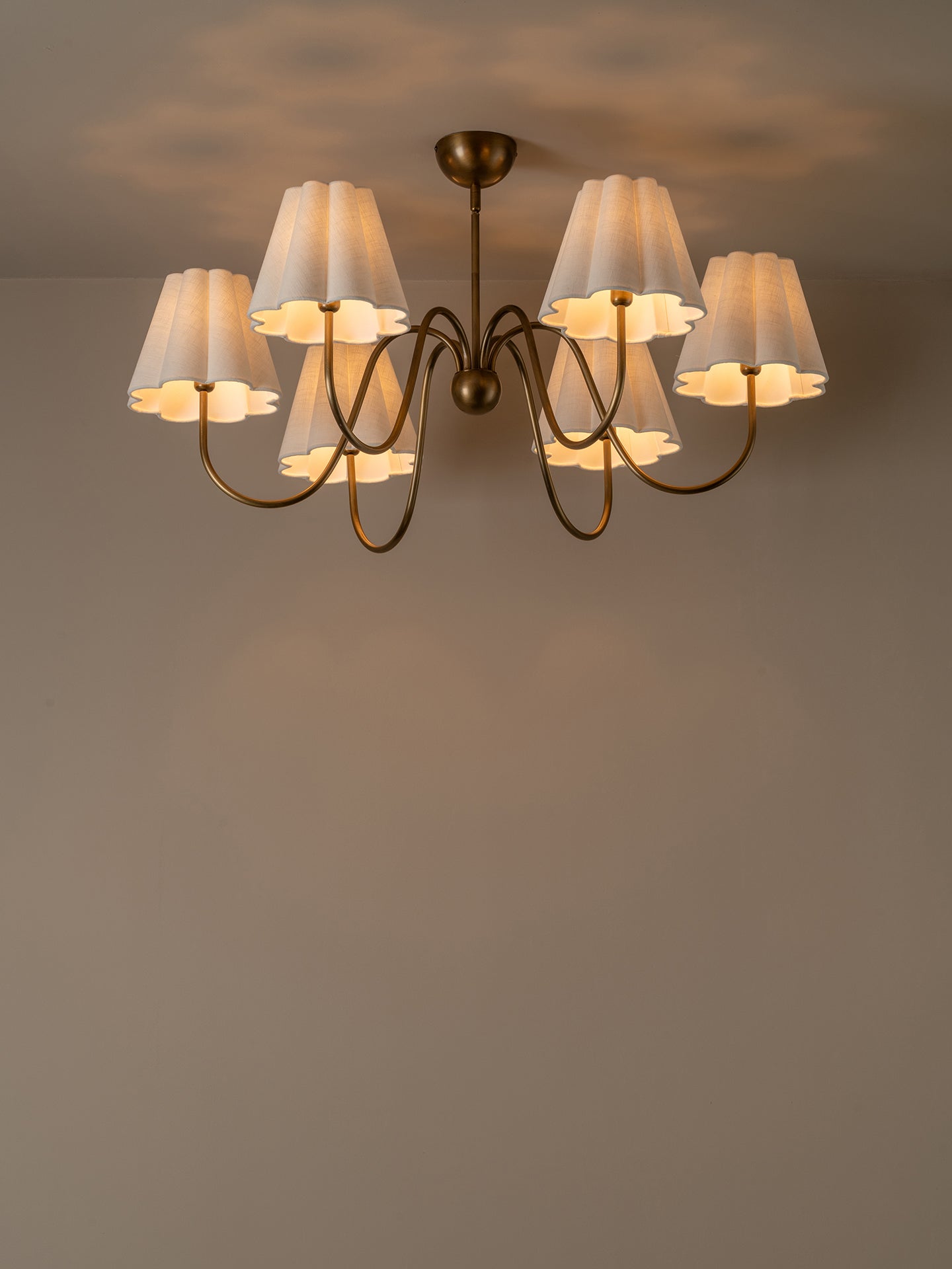 A six-light chandelier with a dark bronze metal frame and pleated white fabric shades, shown illuminated against a neutral warm gray wall and ceiling.