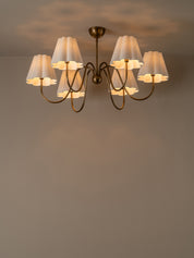 A six-light chandelier with a dark bronze metal frame and pleated white fabric shades, shown illuminated against a neutral warm gray wall and ceiling.
