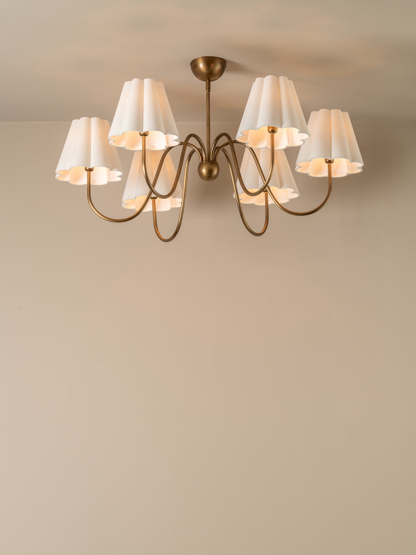A six-light brass chandelier with white pleated drum shades, shown hanging against a plain beige wall and ceiling.
