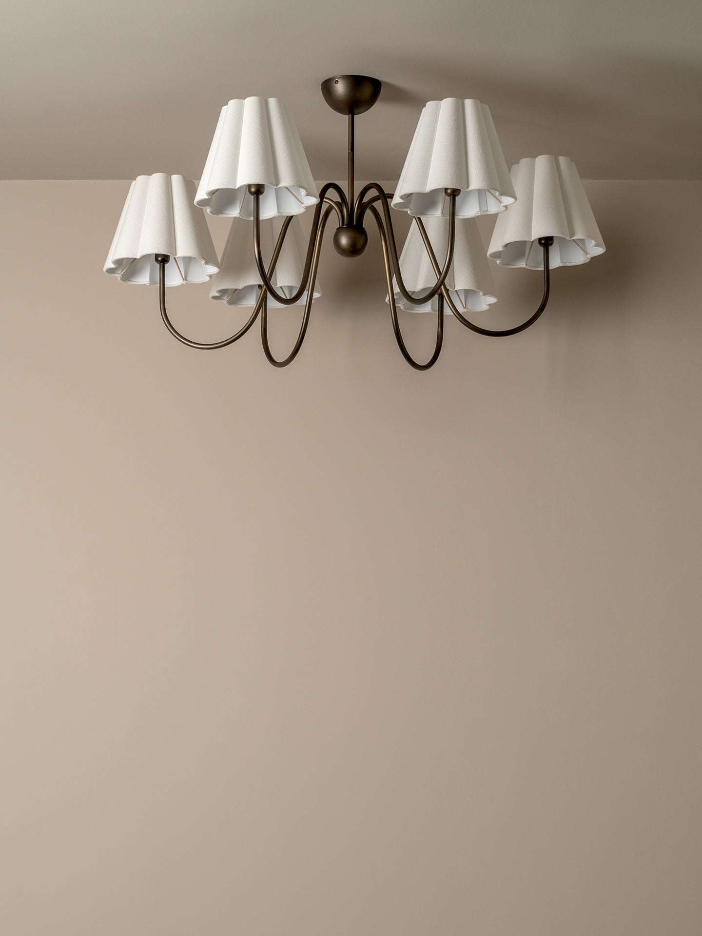A six-light chandelier with white pleated drum shades and a dark bronze metal frame, shown mounted on a beige wall and ceiling.