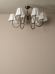 A six-light chandelier with white pleated drum shades and a dark bronze metal frame, shown mounted on a beige wall and ceiling.
