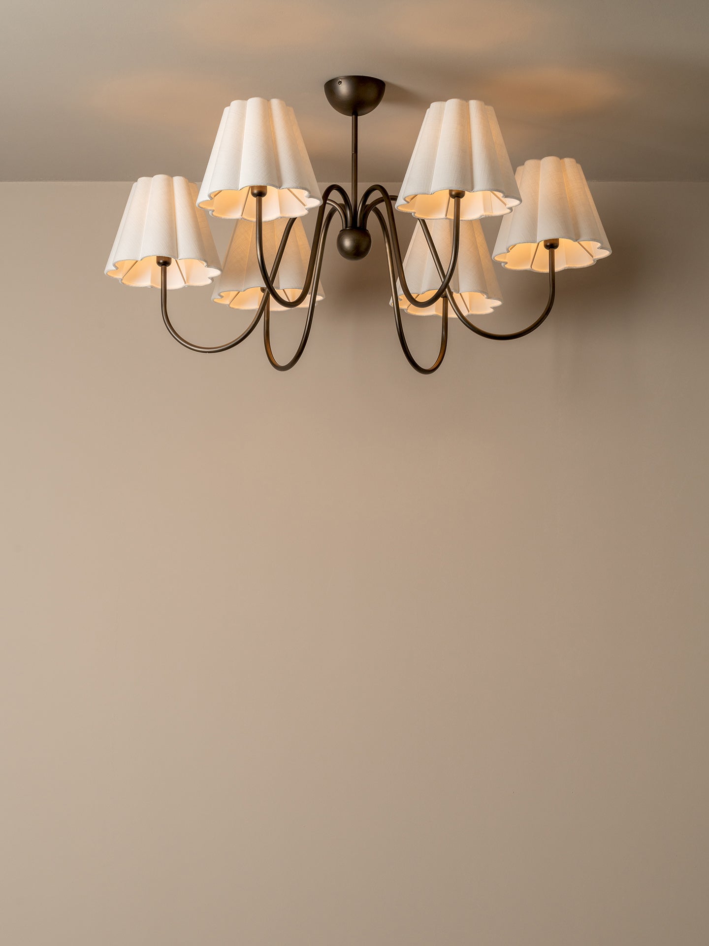 A six-light chandelier with white pleated drum shades and a dark metal frame, shown mounted on a ceiling against a beige wall.