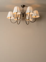 A six-light chandelier with white pleated drum shades and a dark metal frame, shown mounted on a ceiling against a beige wall.