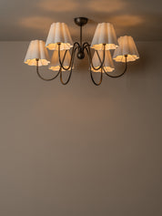 A six-light chandelier with white pleated drum shades and a dark metal frame, shown hanging against a plain wall with warm light glowing from the bulbs.