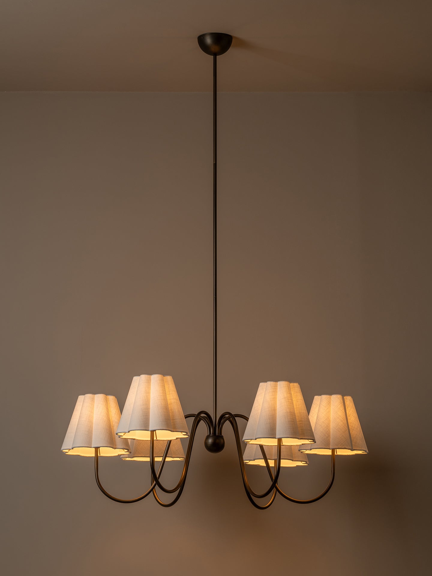 A six-light chandelier with a dark metal frame and pleated off-white fabric shades, illuminated against a warm neutral wall.