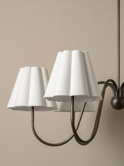 A close-up of a wall-mounted multi-arm light fixture with three white pleated fabric shades and a dark bronze metal frame against a warm beige wall.