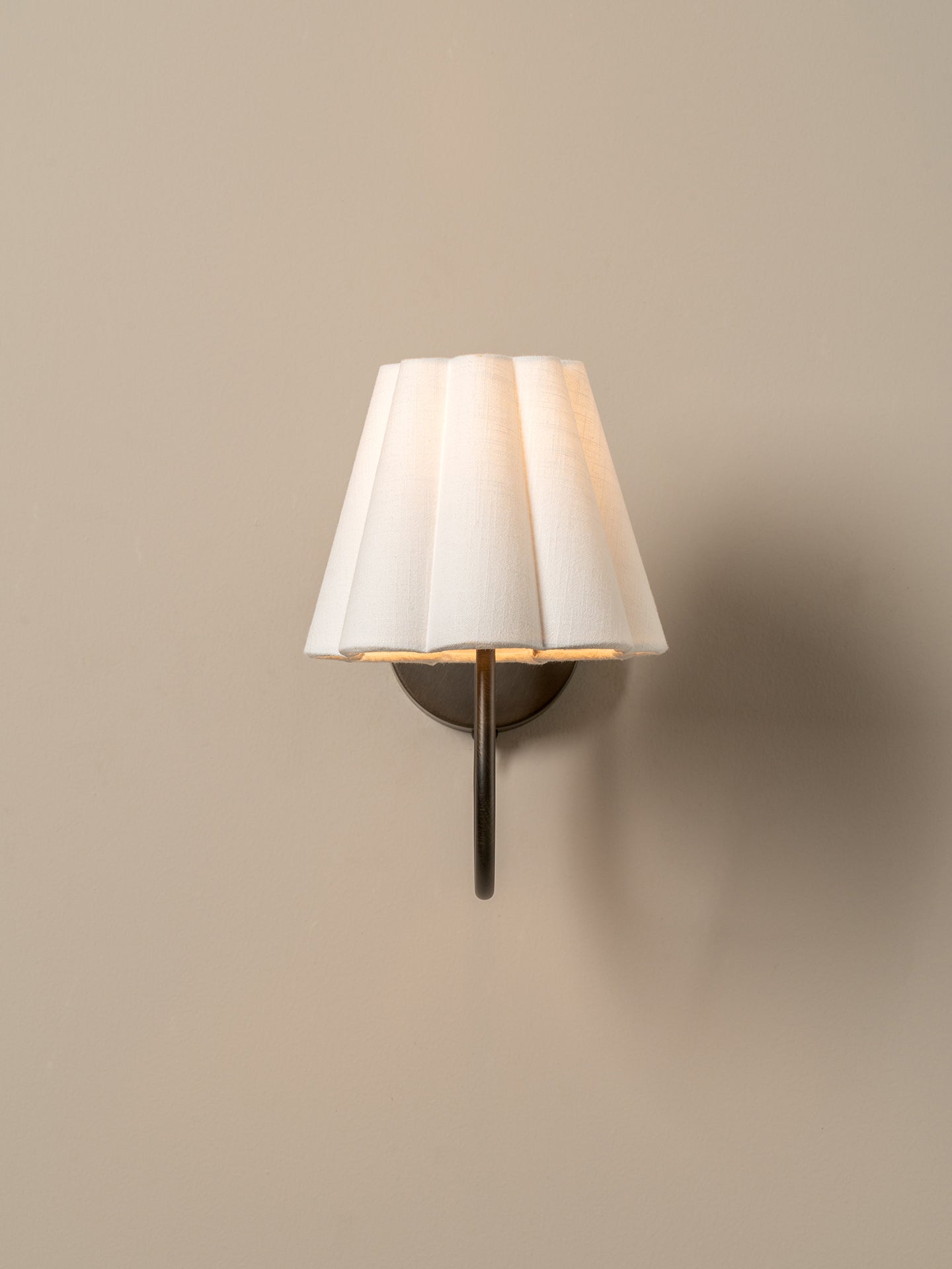 A modern wall-mounted sconce with a pleated white fabric shade and a dark metal stem, shown lit against a plain beige wall.