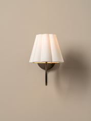 A modern wall-mounted sconce with a pleated white fabric shade and a dark metal stem, shown lit against a plain beige wall.